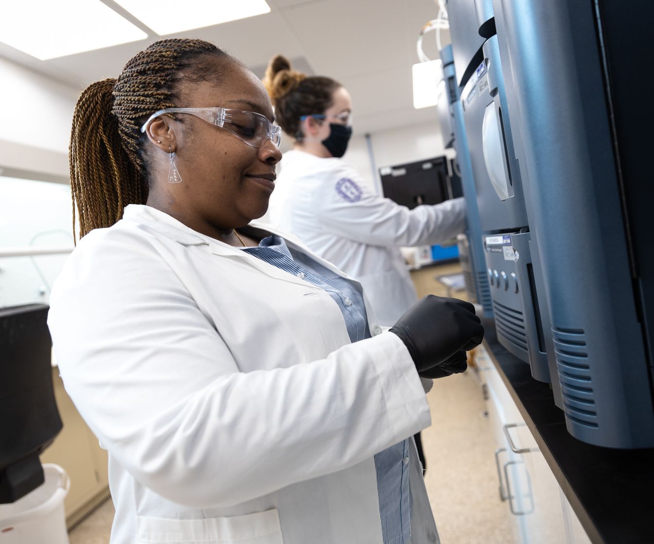 woman in white coat, blue gloves, and eye protection opening a drawer